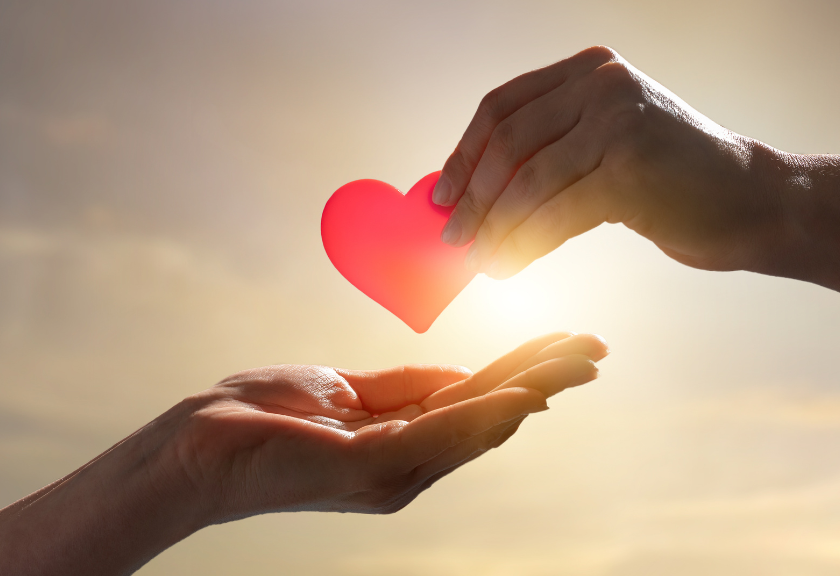 Two hands pass a paper cutout of a heart against a bright sunny sky, mood is caring
